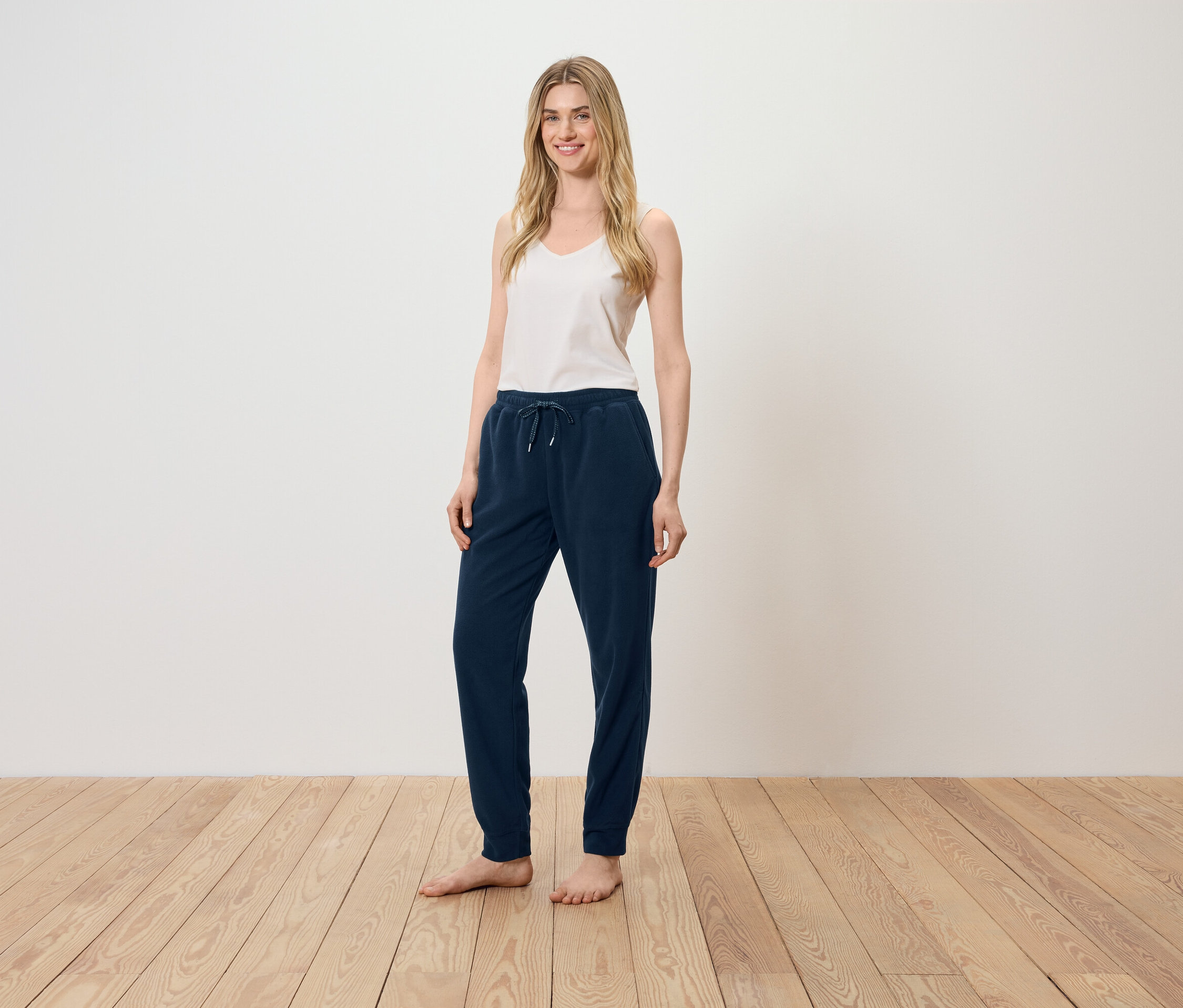 Une femme se tient sur un plancher en bois, portant un débardeur blanc et un pantalon de survêtement bleu marine.