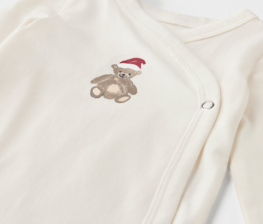 Gros plan sur un body de bébé blanc avec un ours en peluche marron coiffé d'un bonnet rouge.