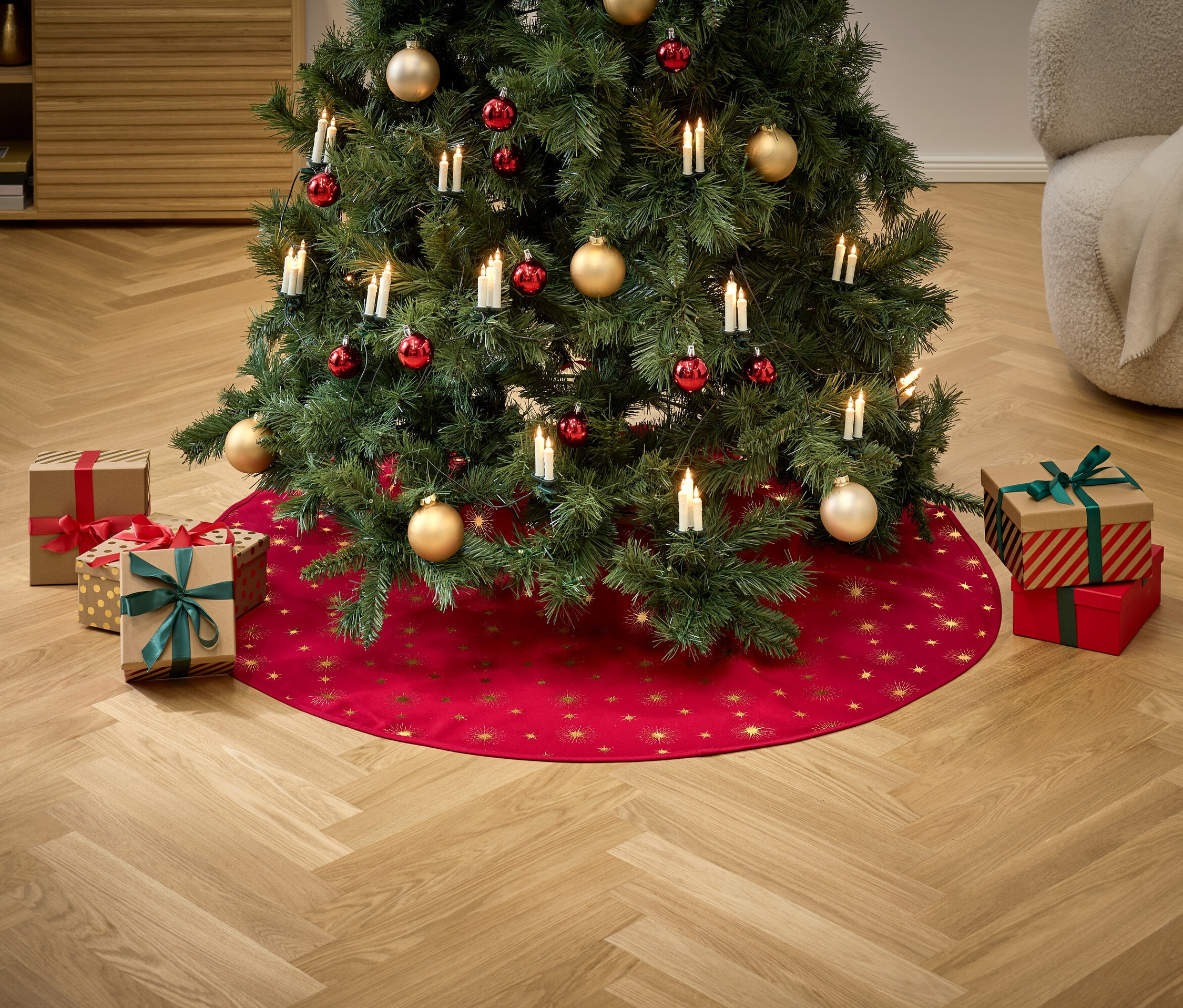 Un arbre de Noël décoré d'ornements rouges et dorés et de lumières se dresse sur une jupe rouge avec des étoiles dorées, entouré de cadeaux.