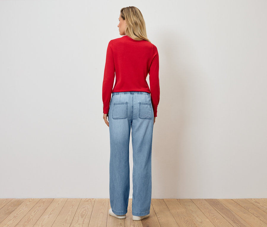 Une femme se tient dos à la caméra portant un pull rouge et un jean bleu.