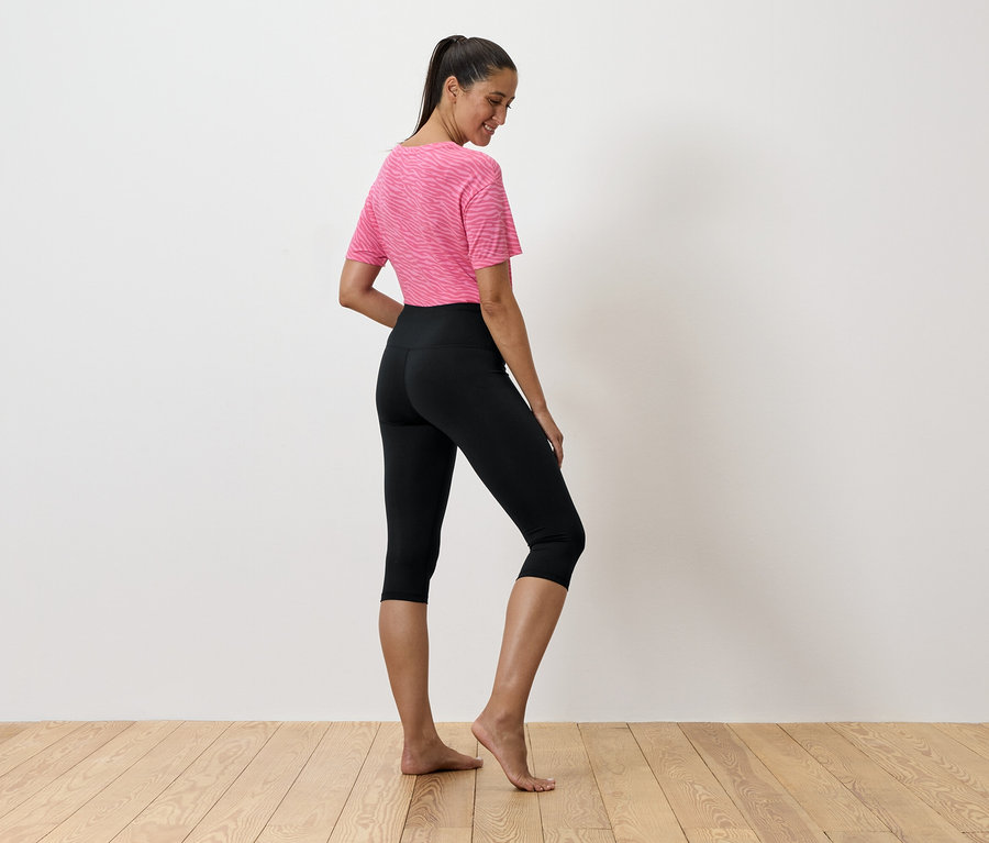 Une femme en T-shirt rose et leggings noirs se tient sur un plancher en bois.