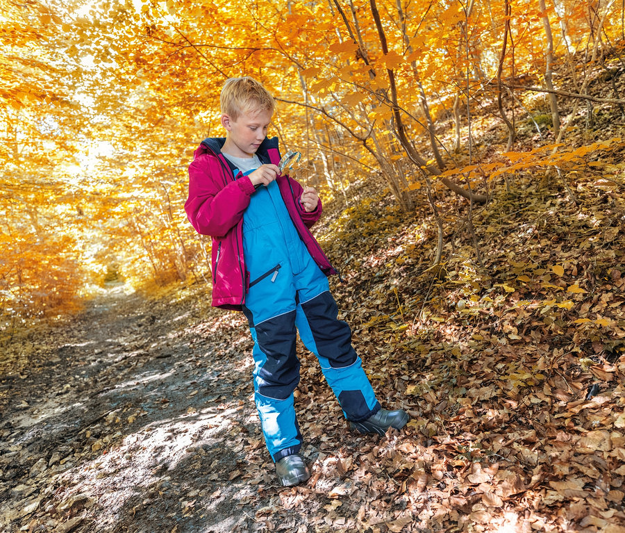 Un garçon avec une veste rouge et un pantalon bleu se tient sur un chemin dans une forêt d'automne et regarde une feuille.