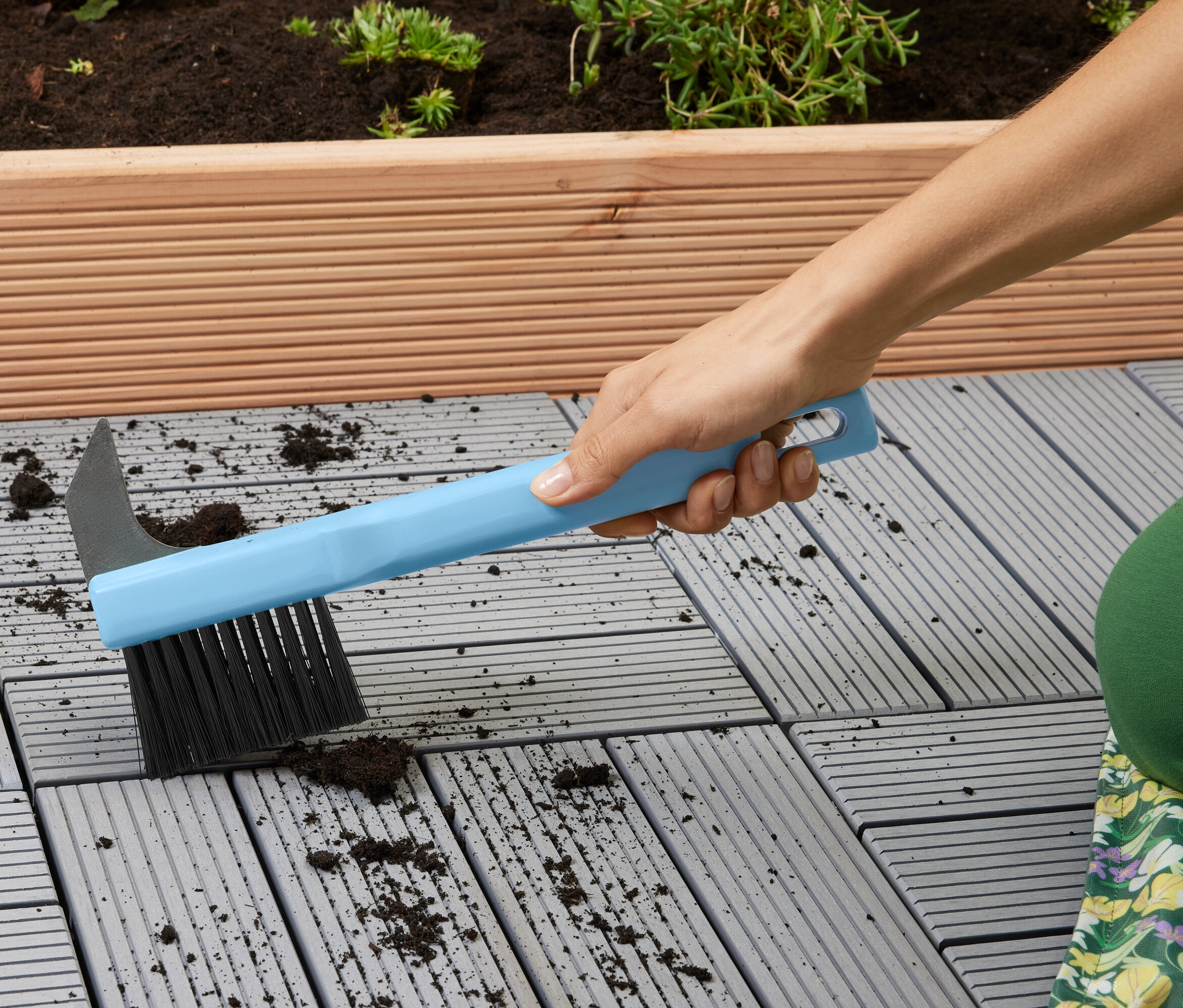 Une femme nettoie des dalles de terrasse grises avec une brosse bleue.