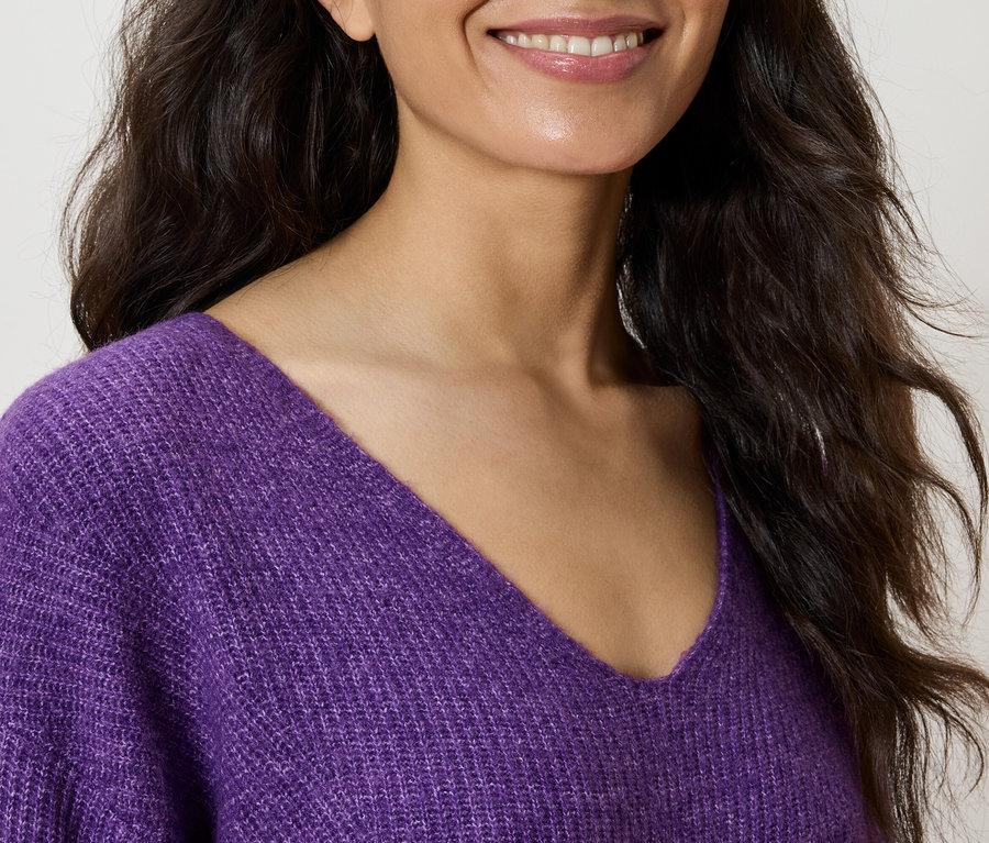 Une femme souriante porte un pull violet à col en V.