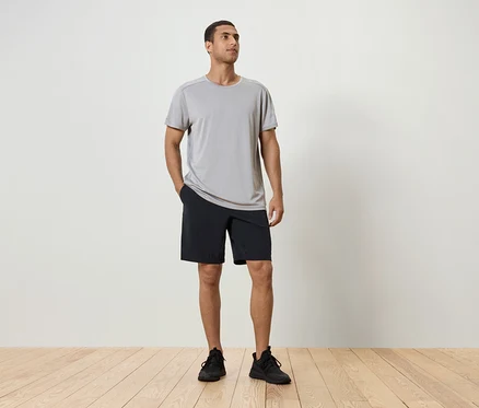 Un homme debout, la main dans la poche de son short noir, portant un t-shirt gris et des chaussures noires.