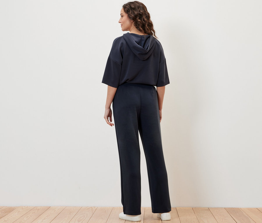 Une femme pose de dos en Joggpants en mélange avec modal et un sweat à capuche devant un mur crème.