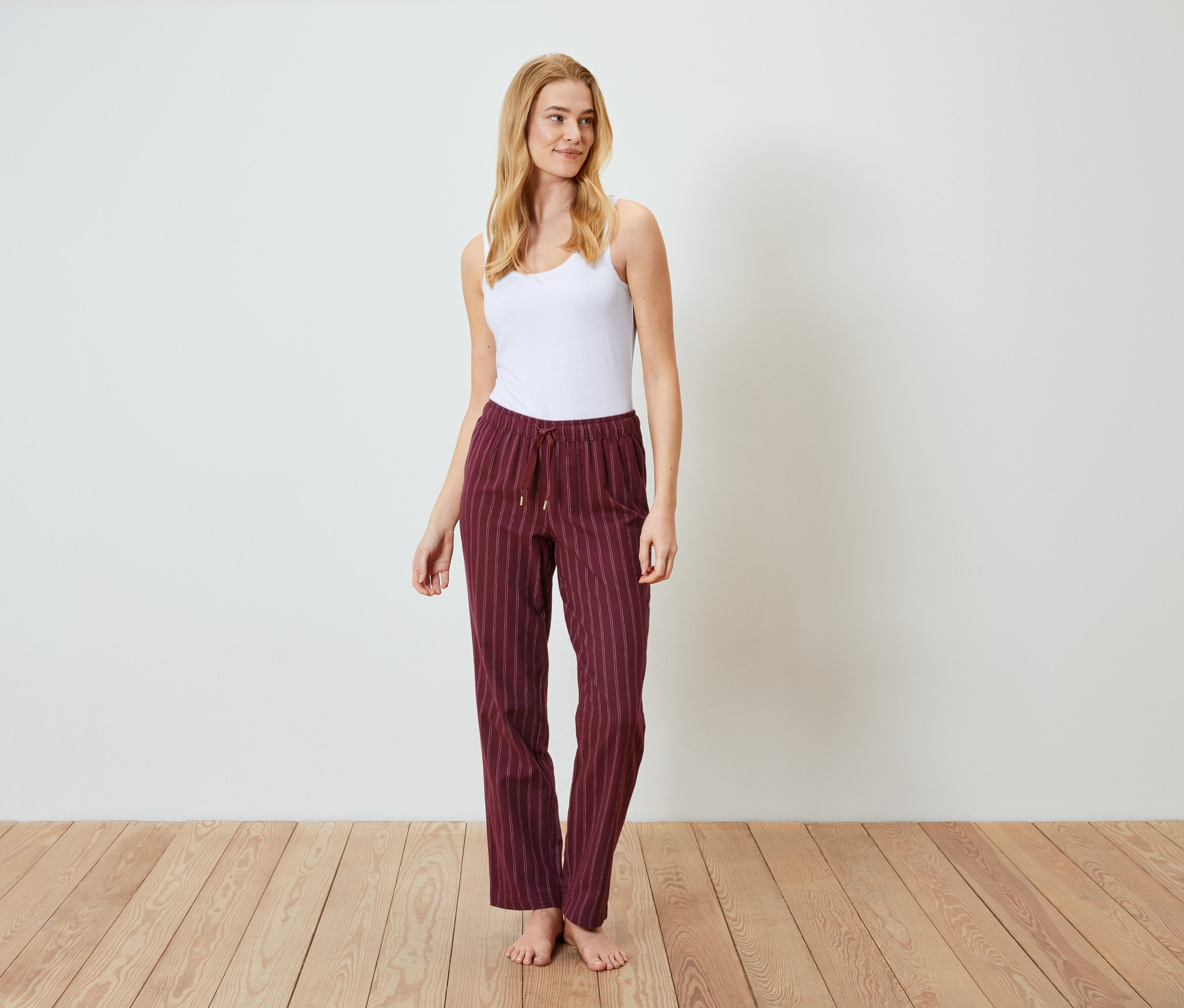 Une femme blonde se tient sur un plancher en bois, portant un débardeur blanc et un pantalon rayé bordeaux.