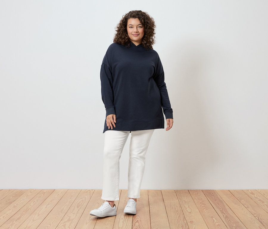 Une femme aux cheveux bouclés se tient devant un mur, portant un sweat à capuche bleu marine, un jean blanc et des baskets blanches.