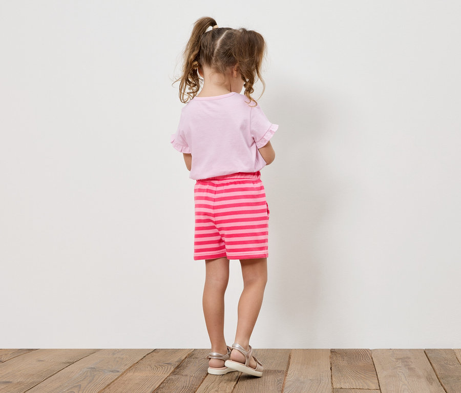 Fillette avec des tresses portant un t-shirt rose et un short rayé, vue de dos.