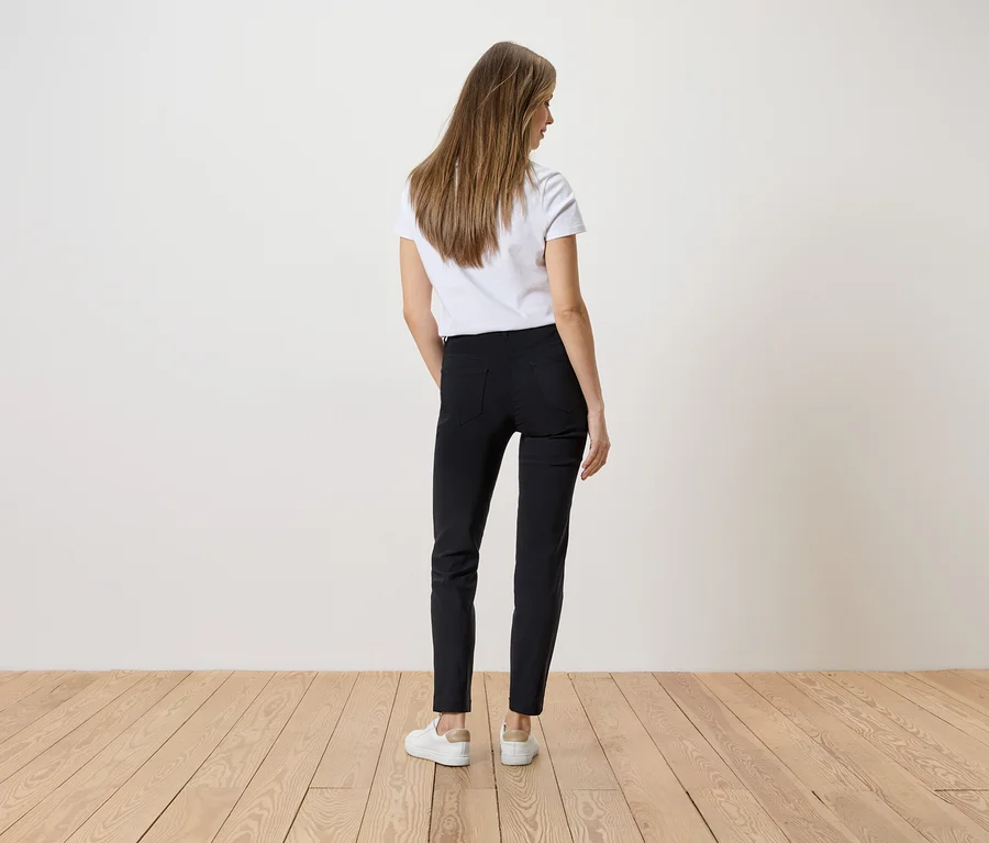 Femme portant un t-shirt blanc et un pantalon noir, vue de dos, sur un plancher en bois.