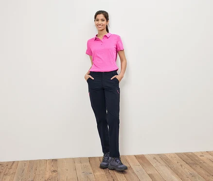 Une femme portant un polo rose et un pantalon foncé se tient devant un mur blanc.