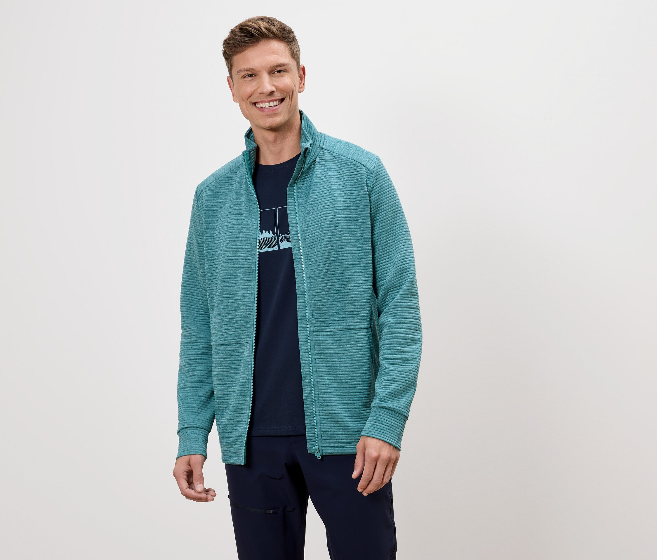 Un homme souriant porte une veste en polaire turquoise sur un t-shirt bleu marine.