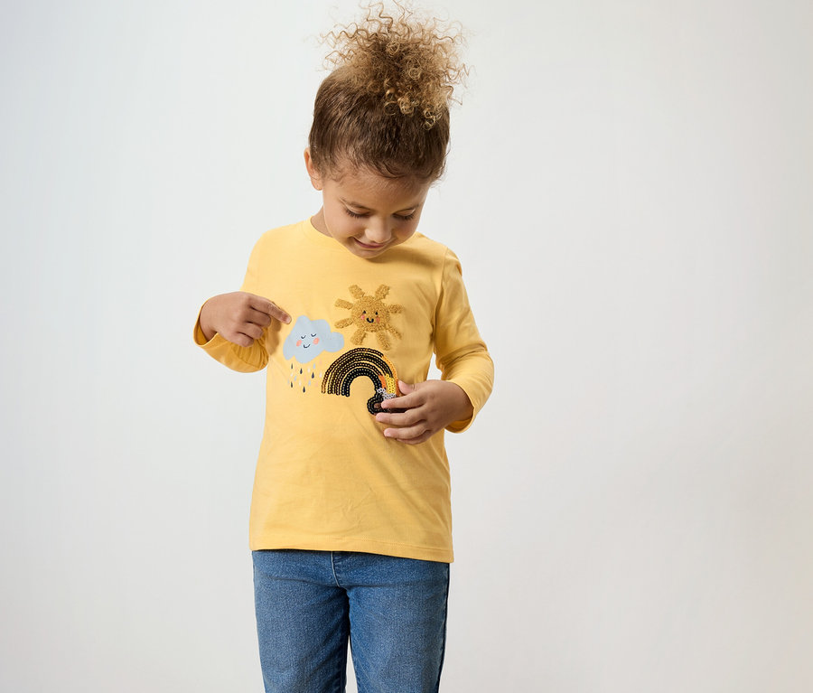 Une jeune fille aux cheveux bouclés regarde son t-shirt jaune à manches longues avec des motifs cousus de nuage, de soleil et d'arc-en-ciel.