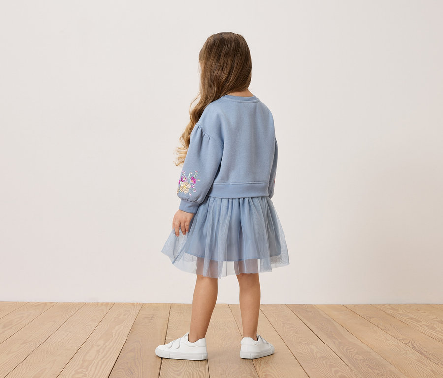 Vue de dos d'une petite fille aux longs cheveux bruns, portant un sweat-shirt bleu avec un motif floral sur la manche et une jupe en tulle assortie, ainsi que des baskets blanches.