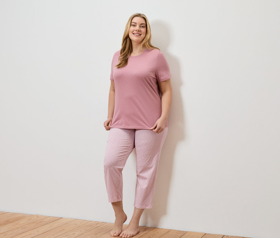 Une femme blonde aux cheveux longs porte un t-shirt rose et un pantalon rose à motifs géométriques.