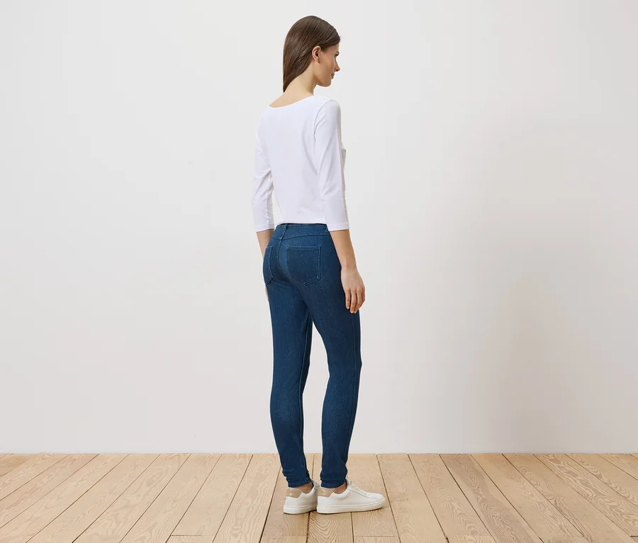 Une femme se tient de profil en jean, haut blanc et chaussures blanches devant un mur blanc et un plancher en bois.