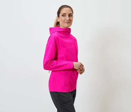 Une femme portant une veste à capuche rose et un pantalon noir pose devant un fond blanc.