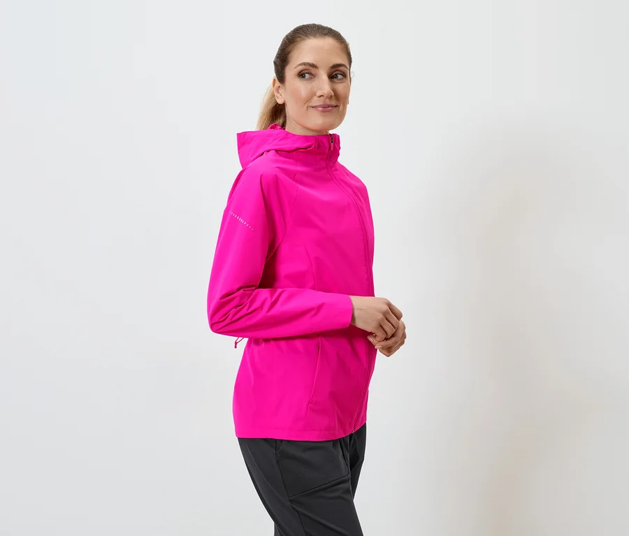 Une femme portant une veste à capuche rose et un pantalon noir pose devant un fond blanc.