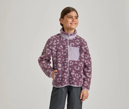 Une jeune fille sourit, regardant vers la droite, vêtue d'une veste polaire à imprimé léopard violet et violet clair avec une poche poitrine zippée et un pantalon gris foncé. Sa main gauche est dans la poche de sa veste.