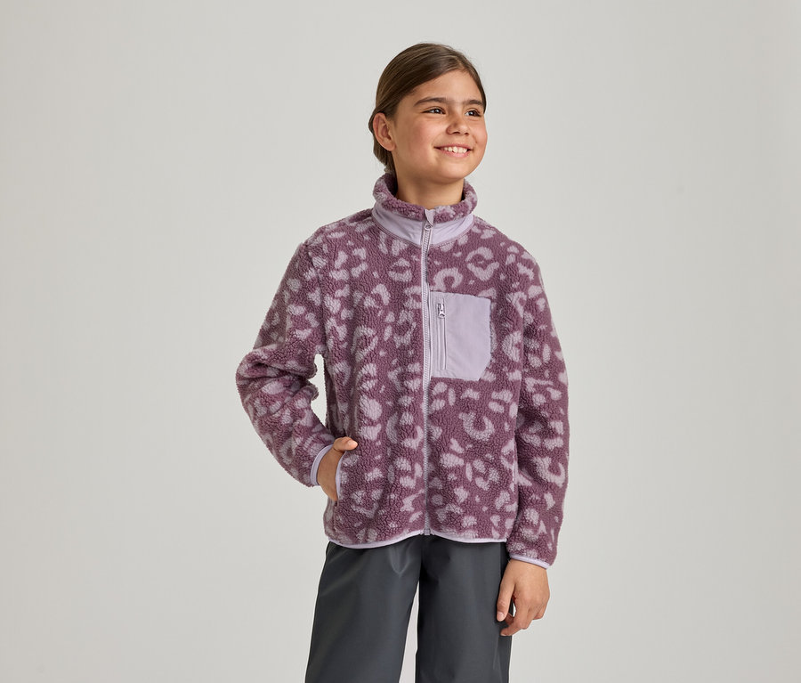 Une jeune fille sourit, regardant vers la droite, vêtue d'une veste polaire à imprimé léopard violet et violet clair avec une poche poitrine zippée et un pantalon gris foncé. Sa main gauche est dans la poche de sa veste.