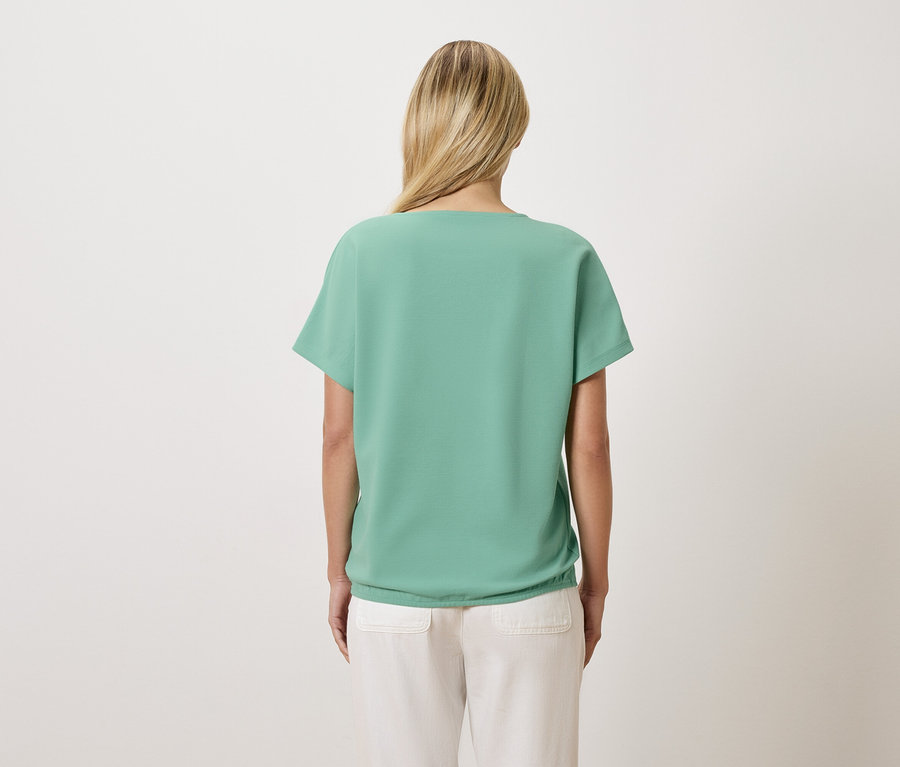 Vue de dos d'une femme aux cheveux blonds portant un t-shirt vert clair et un pantalon clair.