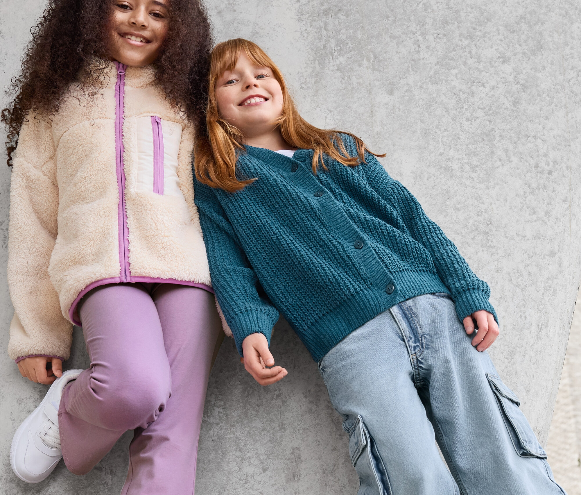 Deux filles sont appuyées contre un mur en béton. La fille de gauche porte une veste en teddy, un pantalon violet et des baskets blanches. La fille de droite porte un cardigan vert et un pantalon bleu.
