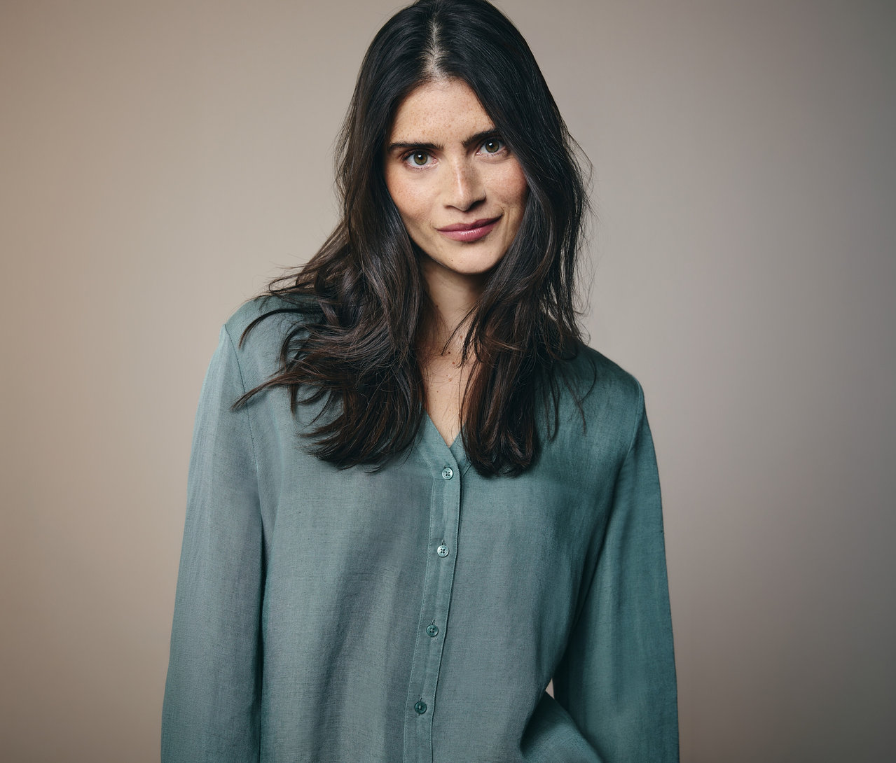 Une femme aux longs cheveux foncés et aux taches de rousseur porte une chemise boutonée bleu-vert sur un fond marron clair.