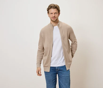 Un homme portant un cardigan zippé marron clair, un t-shirt blanc et un jean sur un fond clair.