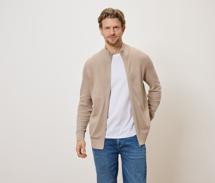 Un homme portant un cardigan zippé marron clair, un t-shirt blanc et un jean sur un fond clair.