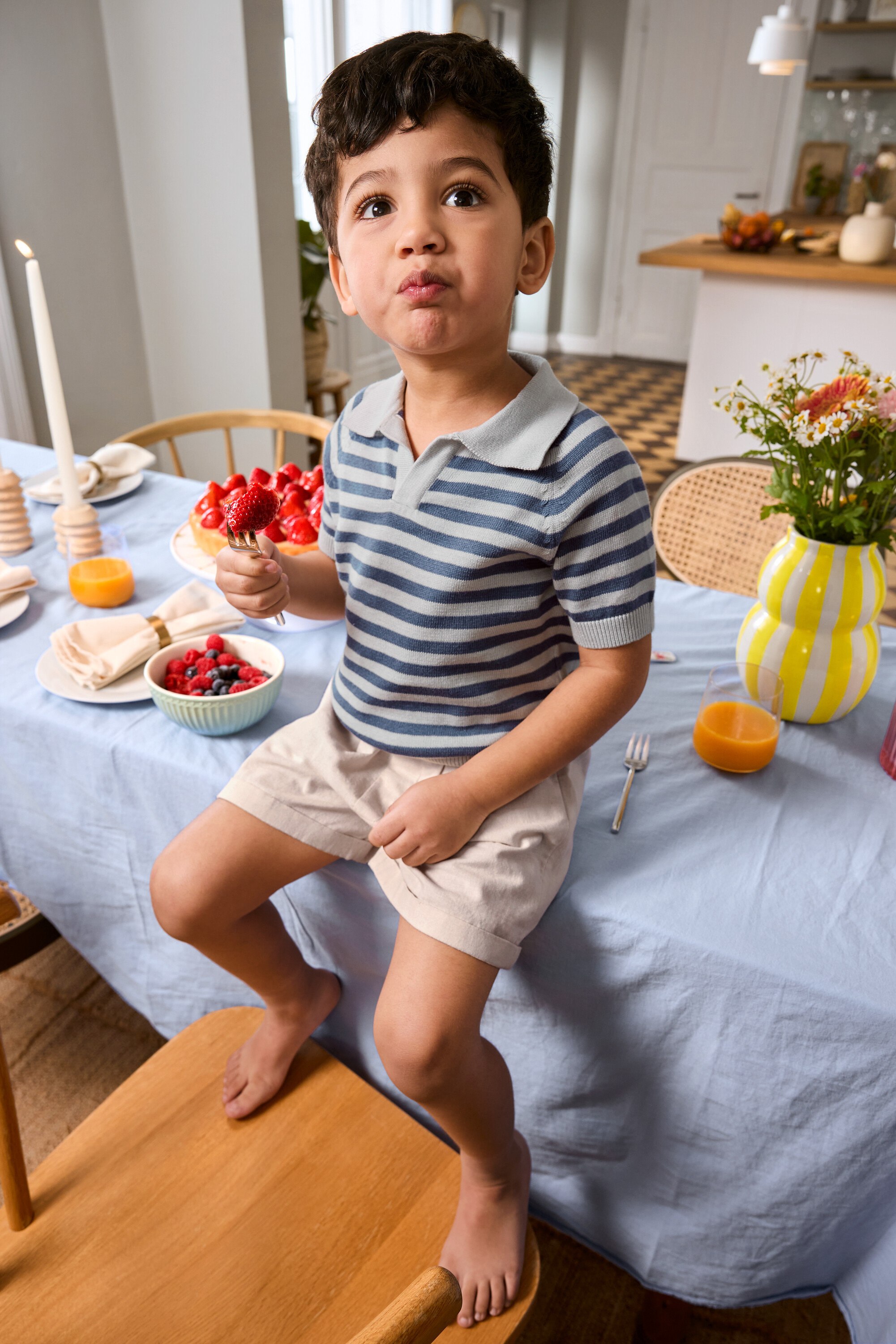 Un garçon mange une fraise sur une fourchette assis sur le bord d'une table, portant un Kinder-Shorts et un Kinder-Strick-Poloshirt. Vase et plateau à tarte sur la table.
