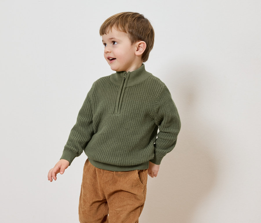 Un garçon aux cheveux bruns porte un pull en tricot vert olive avec une fermeture éclair et un pantalon en velours côtelé marron sur un fond blanc.
