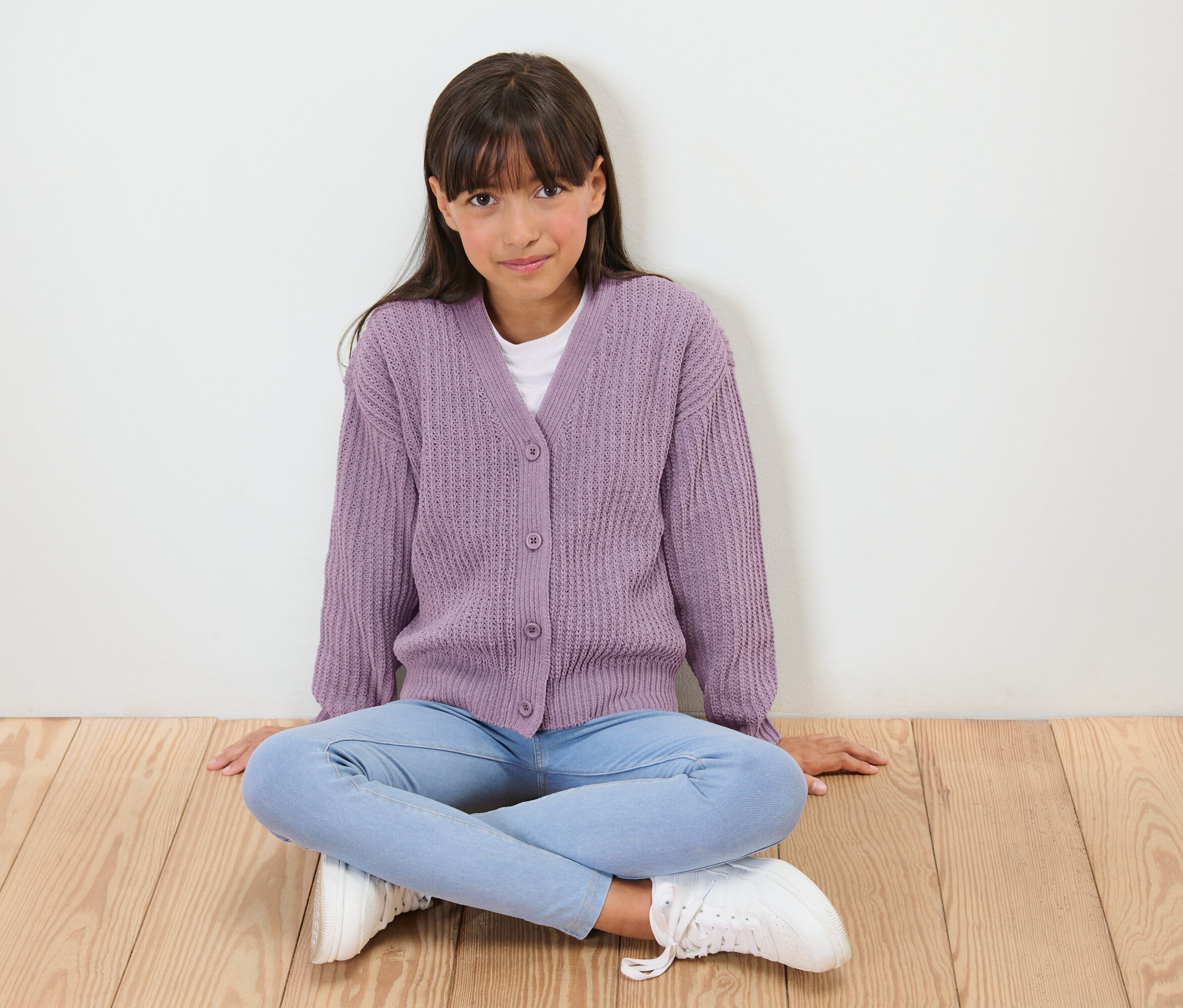 Une fille assise en tailleur sur un plancher en bois, portant un cardigan violet, un jean bleu et des baskets blanches.