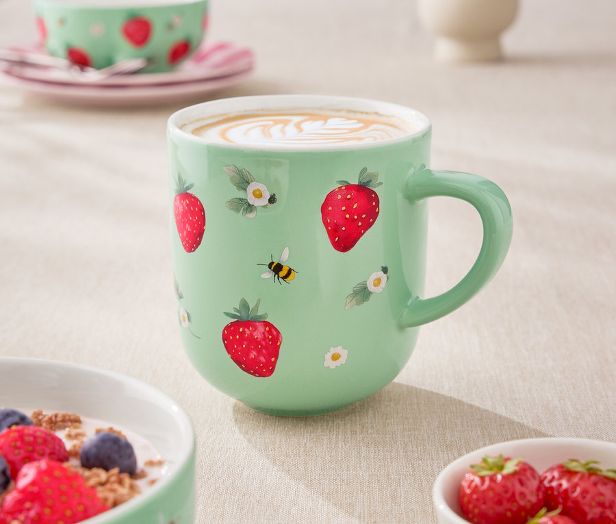 Gros plan d'une table avec une tasse de café vert clair, un bol de céréales avec du yaourt et des baies, et un petit bol de fraises. La tasse est décorée de fraises et de petites fleurs.