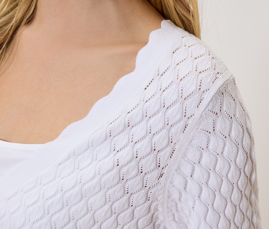 Gros plan d'une femme aux cheveux blonds portant un cardigan tricoté blanc ajouré sur un haut blanc.