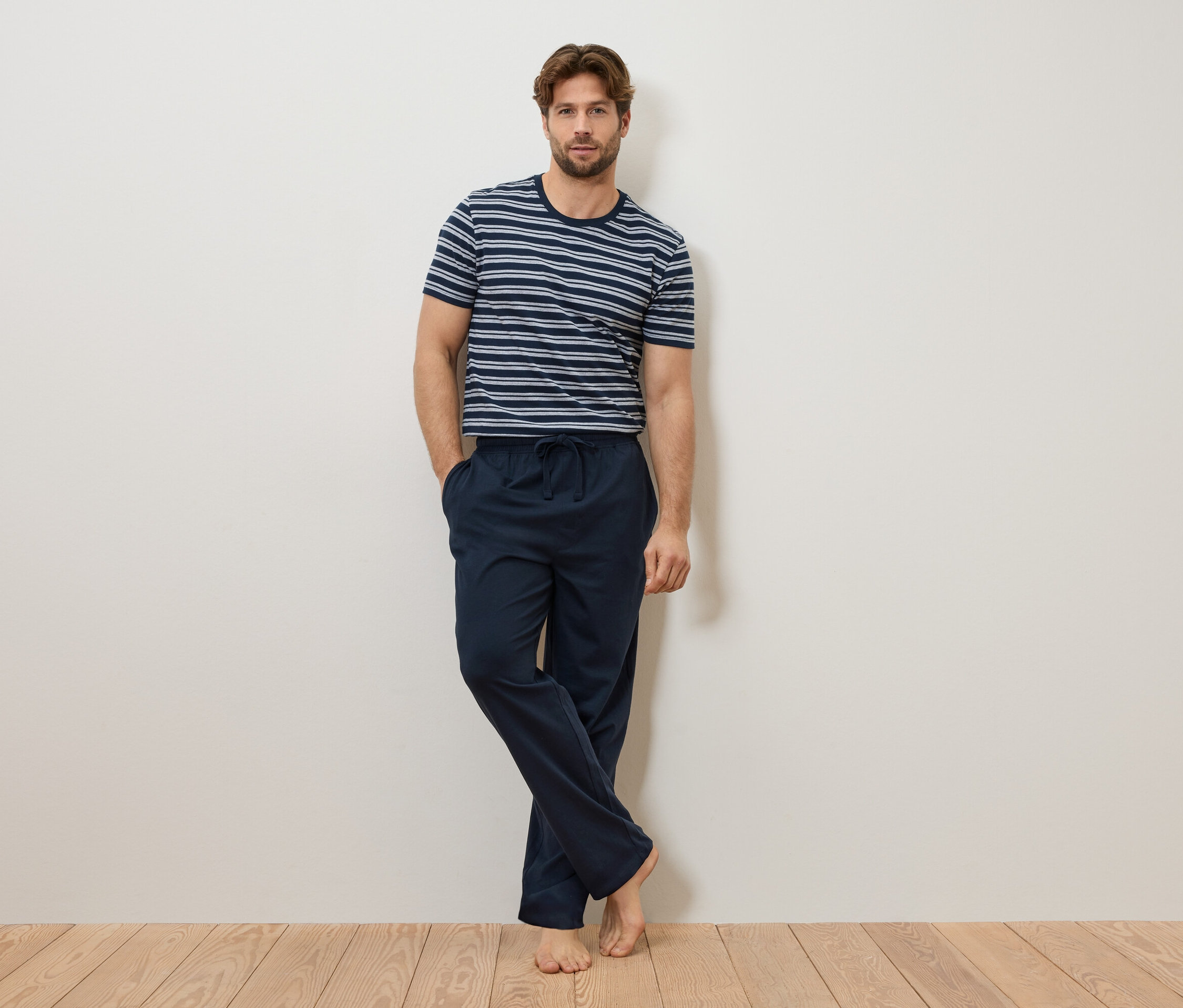 Un homme est appuyé contre un mur, portant un t-shirt rayé bleu et blanc et un pantalon de pyjama bleu marine.