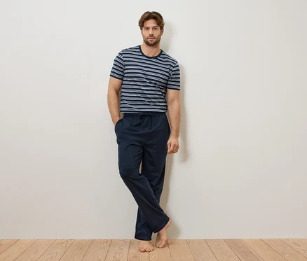 Un homme est appuyé contre un mur, portant un t-shirt rayé bleu et blanc et un pantalon de pyjama bleu marine.