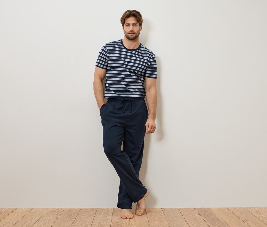 Un homme est appuyé contre un mur, portant un t-shirt rayé bleu et blanc et un pantalon de pyjama bleu marine.