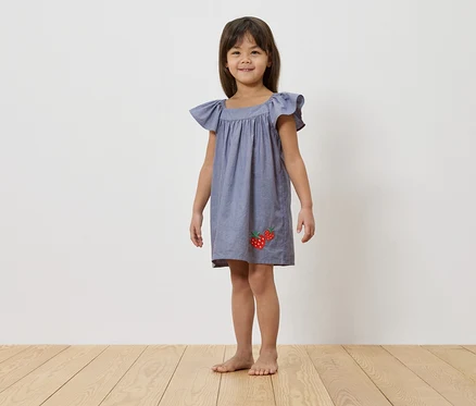 Une jeune fille se tient pieds nus sur un plancher en bois devant un mur blanc, portant une robe bleue avec des appliqués de fraises.