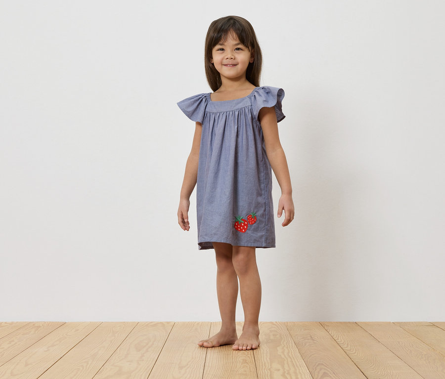 Une jeune fille se tient pieds nus sur un plancher en bois devant un mur blanc, portant une robe bleue avec des appliqués de fraises.
