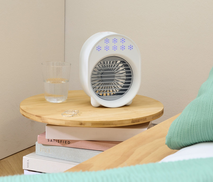 Un ventilateur portable blanc avec des flocons de neige bleus est posé sur une petite table en bois à côté d'un verre d'eau et de bagues.