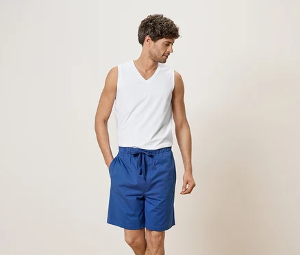 Homme portant un débardeur blanc et un short bleu, la main dans la poche.