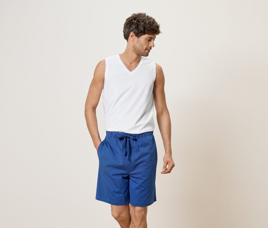 Homme portant un débardeur blanc et un short bleu, la main dans la poche.