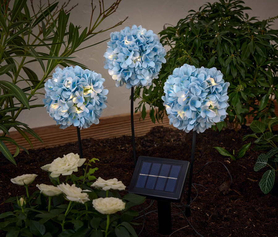 Trois piques solaires «
Hortensia
» illuminent des roses blanches et des plantes dans un parterre.