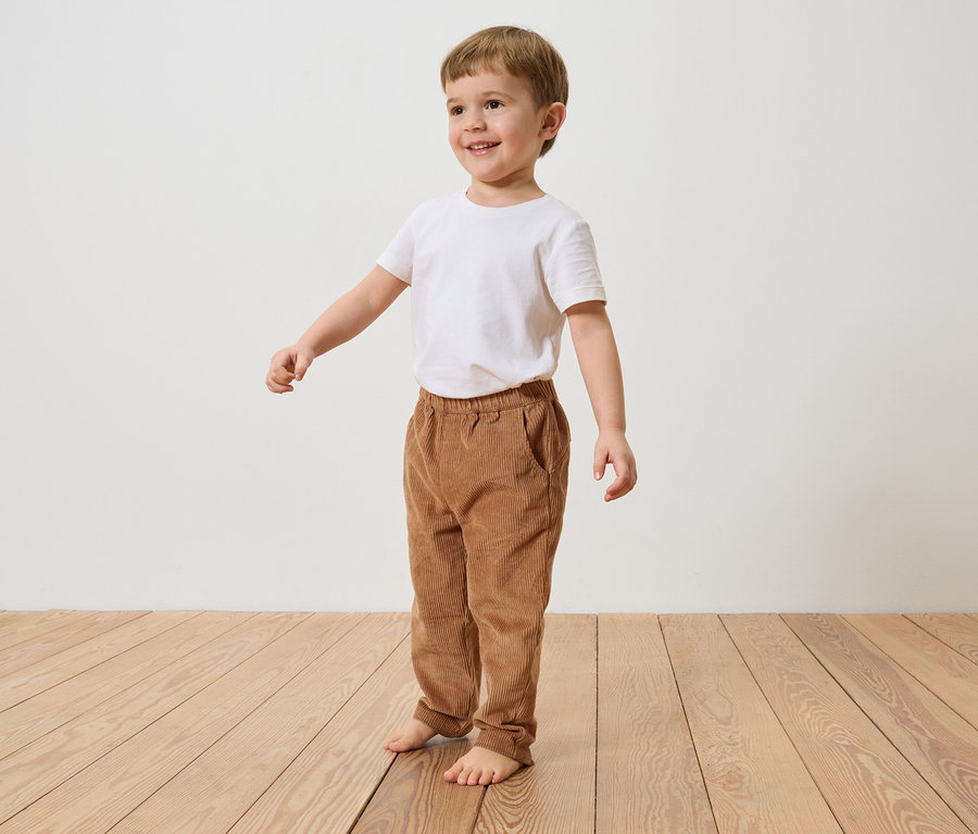 Un garçon souriant aux cheveux bruns se tient pieds nus sur un plancher en bois, vêtu d'un t-shirt blanc et d'un pantalon en velours côtelé marron.