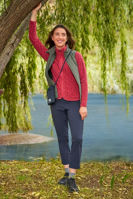 Une femme s'appuie contre un arbre au bord d'un lac, portant une veste en polaire rouge, un pantalon bleu marine et un gilet kaki »packable« Hybrid-Weste.