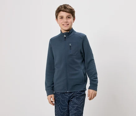 Un garçon aux cheveux bruns porte une veste bleu marine zippée et un pantalon à motifs bleus sur fond blanc.
