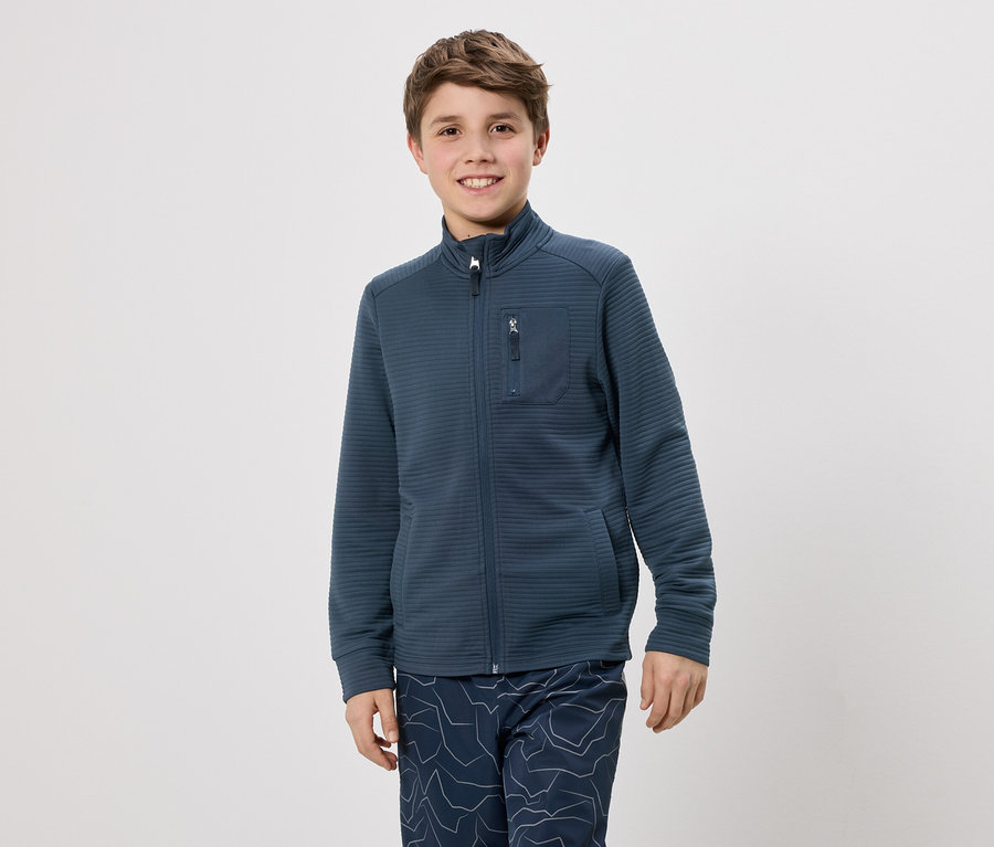 Un garçon aux cheveux bruns porte une veste bleu marine zippée et un pantalon à motifs bleus sur fond blanc.