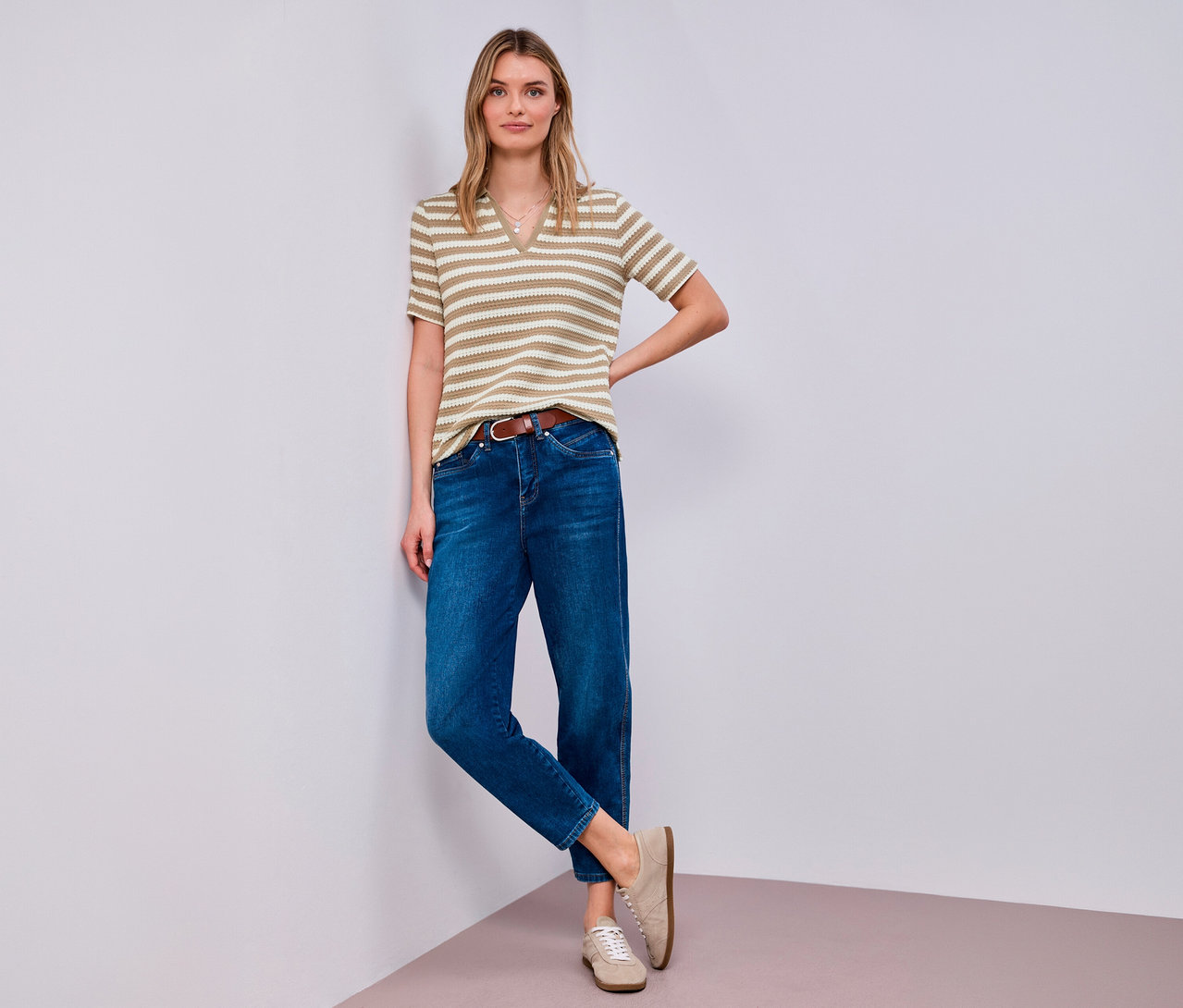 Une femme pose dans un coin avec un pull en maille Street One, un jean, une ceinture marron et des chaussures beiges.