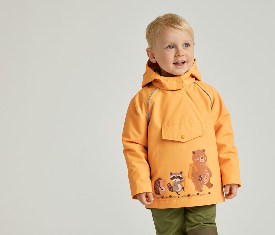 Un petit garçon aux cheveux blonds courts porte une veste orange avec un motif d'ours, de raton laveur et de hérisson, et un pantalon vert.