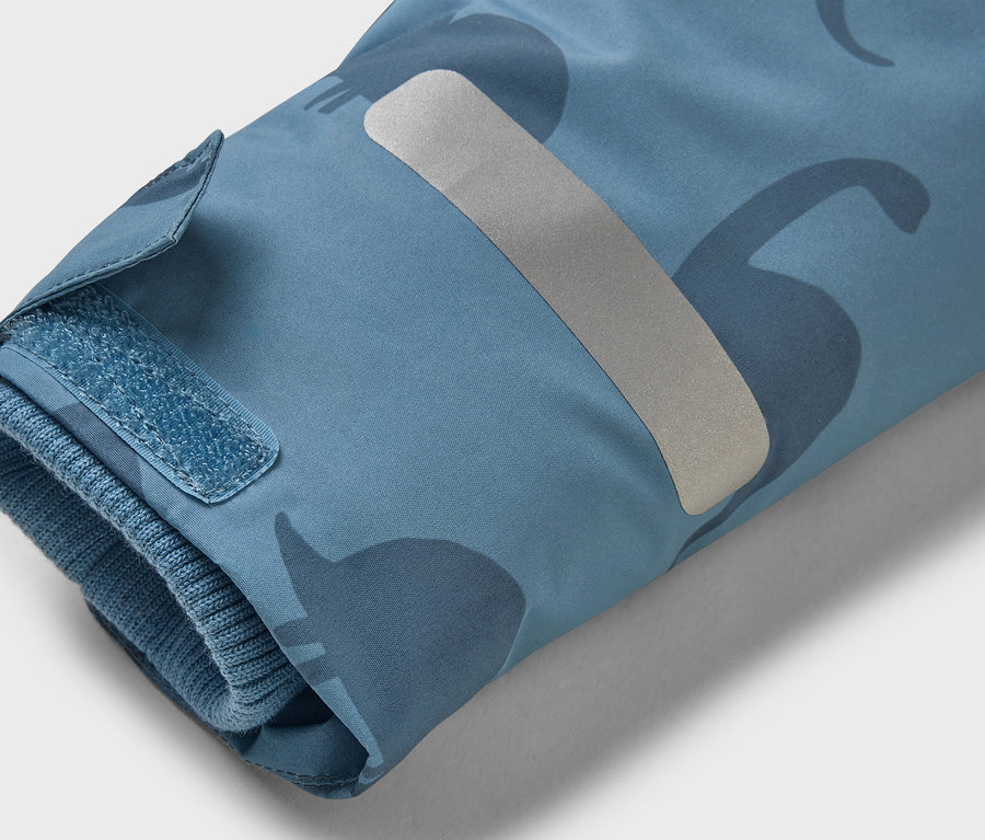 Gros plan sur la manche d'une veste bleu clair à motif de dinosaures, avec une fermeture velcro et une bande réfléchissante.